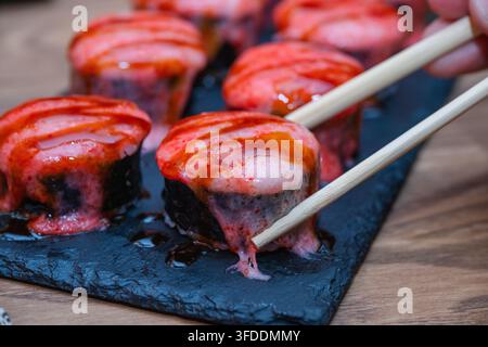 Ein Paar Essstäbchen hält eine einzelne gebackene Sushirolle, die mit rosa Sauce und süßer Sozenglasur überzogen ist. Stockfoto