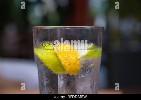 Ein mit Kondenswasser bedecktes Glas, gefüllt mit einem kalten Getränk, das Scheiben von Zitrone und Limette enthält, steht vor einem verschwommenen Hintergrund. Stockfoto