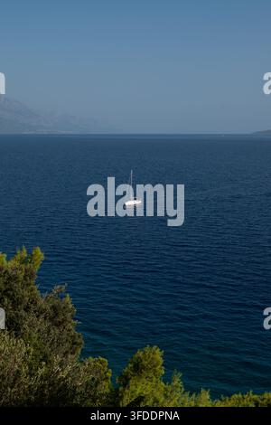 Einsames Segelboot treibt über ruhiges blaues Meer unter dunstigen Bergen Stockfoto