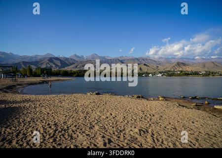 Eine Weitwinkel-Panoramadarstellung zeigt das sandige Ufer des Issyk-Kul-Sees, mit einer Person, die im klaren blauen Wasser watet, und einer entfernten Stadt, Cholpon-Ata, Stockfoto