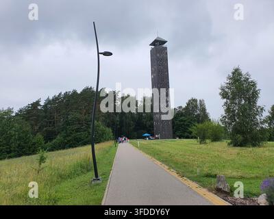 Ein hoher hölzerner Aussichtsturm mit geometrischen Ausschnittmustern steht markant vor einem bewölkten Himmel, gesehen von einem gepflasterten Weg, der führt Stockfoto