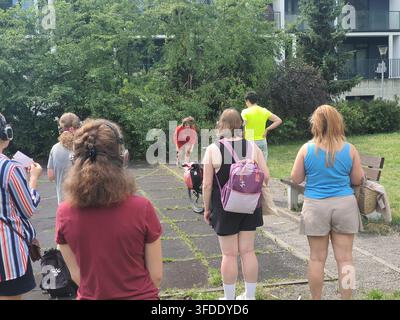 Eine Gruppe von sieben Personen, die im Freien in einer städtischen Parkumgebung versammelt ist und einen Darsteller in roter Kleidung beobachtet, der mit einem Fahrrad interagiert. Die Szene spielt Stockfoto