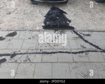 Ein großer schwarzer Öl- oder Flüssigkeitsaustritt auf einem gepflasterten Gehweg und dem angrenzenden Kiesboden, der von unter der vorderen Stoßstange eines geparkten Autos stammt, das am Stockfoto