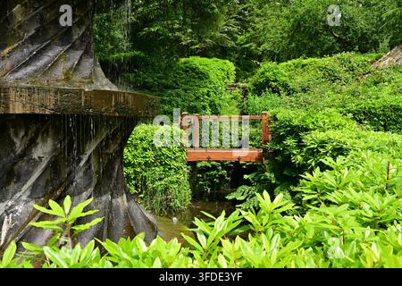 Hölzerne Gartenbrücke und spiralförmiger Wasserfall in üppiger Vegetation Stockfoto