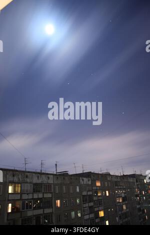 Stadtlandschaft bei Nacht, die einen hellen Vollmond zeigt, der durch Bewegungsunschärfe-Wolken im dunklen Himmel über einem typischen Wohnblockhaus scheint. Stockfoto