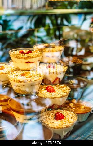 Verschiedene geschichtete Desserts in klaren Plastikbechern, die in einer gekühlten Backwarenvitrine präsentiert werden. Mehrere klare Plastikbecher enthalten verschiedene süße Leckereien, darunter Parfaits mit zerbröckelten Keksen, Sahne und Erdbeeren, Schokoladen-Mousse-Becher und Karamell-Pecan Stockfoto