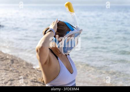 Eine Frau steht an einem kiesigen Ufer und passt eine Schnorchelmaske neben ruhigem Wasser an. Stockfoto