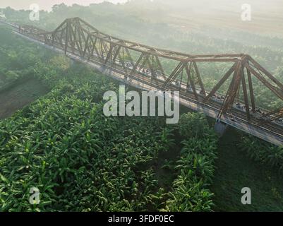 Neblige Luftaufnahme der Vinh-Tuy-Brücke, die sich über eine üppige Bananenplantage in der Nähe von Hanoi, Vietnam, erstreckt, mit Stahlträgern, ländlichem Ackerland und sanftem Morgen Stockfoto