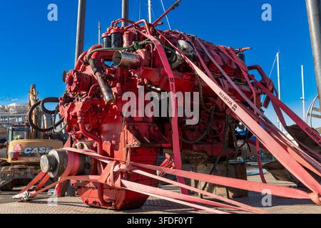 Portimao - Algarve - Portugal - 11-27-2025: Großer roter Motorblock, gesichert mit Hebegurten und Seilen auf einem Dock, mit Booten und Arbeitsfahrzeugen unter klarem blauem Himmel sichtbar. Stockfoto