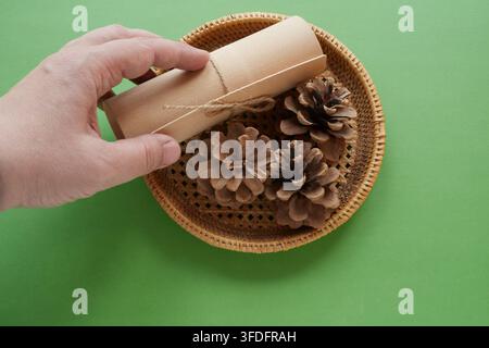 Eine Hand legt Geschenkpapier in einen Weidenkorb mit Tannenzapfen, grüner Hintergrund, Nahaufnahme. Stockfoto
