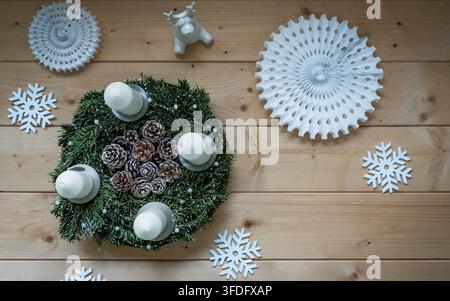 Ansicht von oben eines traditionellen Adventskranzes aus immergrünen Zweigen, verziert mit Tannenzapfen und vier weißen Kerzen auf einem hellen Holzgrund Stockfoto