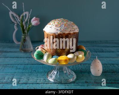 Traditionelle osteuropäische Paska, auch bekannt als Pasca, auf einem gläsernen Kuchenständer präsentiert, umgeben von bunten dekorativen Eiern, Tulpen und Frühlingsdekoration Stockfoto