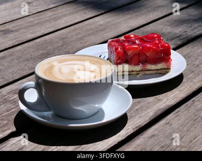 Hochwinkelaufnahme zeigt eine weiße Tasse mit warmem Cappuccino mit detaillierter Latte Art neben einem Stück frischem Erdbeerkuchen mit roter Glasur. Serviert auf Stockfoto