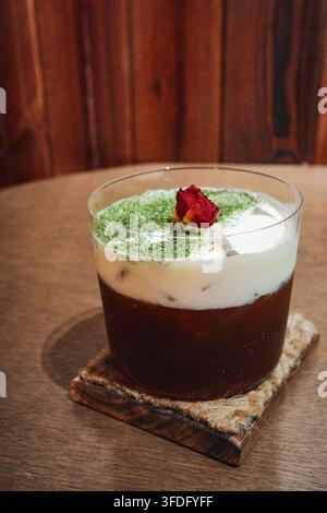 Der dunkelbraune Latte im durchsichtigen Glas ist mit weißem Milchschaum bedeckt. Der Milchschaum ist mit hellem Matcha-Pulver bestreut, und ein dunkelroter Dr. Stockfoto
