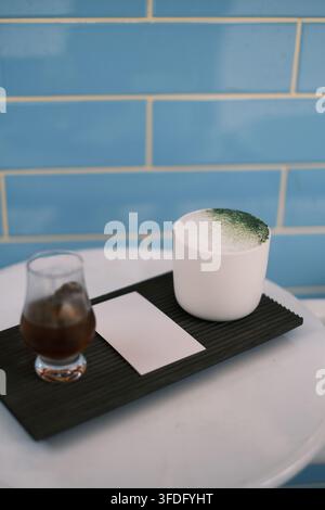 Eine kurze weiße Porzellantasse enthält einen milchig weißen Matcha-Latte, mit smaragdgrünem Matcha-Pulver oben drauf gestreut. Das kleine Glas daneben enthält ein i Stockfoto