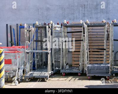 Weite Ansicht, die leere Metallgitterrollenbehälter, rote Einkaufswagen und Holzpaletten zeigt, die draußen an einer grauen Gebäudewand geparkt sind. Das Sonnenlicht c Stockfoto