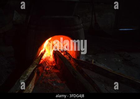 Leuchtende orangefarbene Flammen und glühende Glut in einem traditionellen Lehmofen. Feuerholzscheite brennen in einer dunklen, rustikalen Umgebung, die zum Kochen oder zur Wärme genutzt wird. Stockfoto