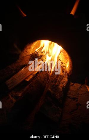 Nahaufnahme eines lodernden Holzfeuers in einem traditionellen Lehmofen. Warme orangefarbene Flammen beleuchten das Brennholz in einer dunklen rustikalen Umgebung. Stockfoto