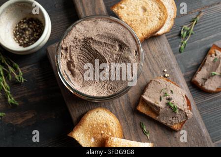 Hausgemachte Hühnerleberpaste im Glas mit geröstetem Brot und Kräutern auf dunklem Holzgrund, Ansicht von oben. Textur der Leberpaste. Stockfoto