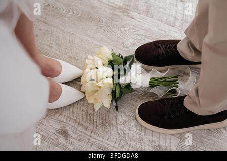 Hochwinkelaufnahme eines Brautstraußes aus weißen Rosen mit grünen Blättern und Satinband, der auf einem hellen Holzboden zwischen den weißen Absätzen der Braut liegt Stockfoto