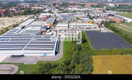 Luftaufnahme einer Industrieanlage mit großen Lagerhallen, die mit Solarmodulen auf dem Dach ausgestattet sind, einer angrenzenden bodengestützten Solarfarm und umgebendem Grün Stockfoto