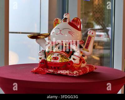 Mittlere Aufnahme einer traditionellen weiß-goldenen Maneki Neko Glückskatzen-Figur, die auf einem rot bezogenen runden Tisch in einer modernen Location ruht, die ein Stockfoto
