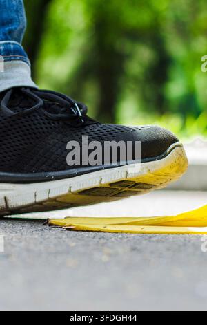 Nahaufnahme eines Mannes Fuß, der einen Sneaker trägt und auf einer Banane auf dem Boden tritt. Die Bananenschale und das zerdrückte Obst erzeugen eine humorvolle Rutschgefahr-Szene Stockfoto