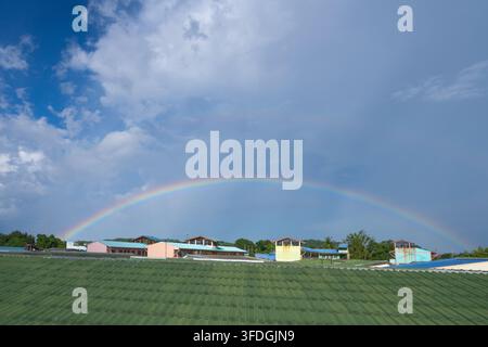 Voller Regenbogen, der sich über den blauen und bewölkten Himmel über grünen Dächern in Thailand nach dem Regen erstreckt. Farbenfrohes natürliches Wetterphänomen während hellem Tro Stockfoto