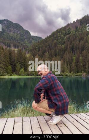 Senior mann, der auf einer Holzterrasse an einem See hockt, umgeben von Bergen und Wald Stockfoto
