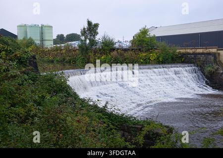 Schräge Ansicht des Warth-Wehrs mit strömendem Weißwasser, eingerahmt von überwucherten Büschen und Industriegebäuden im Hintergrund in Radcliffe, Lancs, UK. Stockfoto