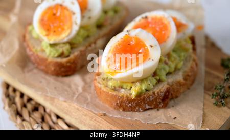 Zwei Scheiben knuspriges Sauerteigbrot mit Avocadopüree und perfekt weich gekochten Eiern mit laufendem Eigelb. Die natürliche Seitenbeleuchtung unterstreicht den Stockfoto