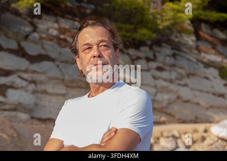 Ein mittelalter Mann in einem weißen Hemd steht im Freien mit verschränkten Armen vor einer sonnenbeschienenen Steinmauer. Stockfoto