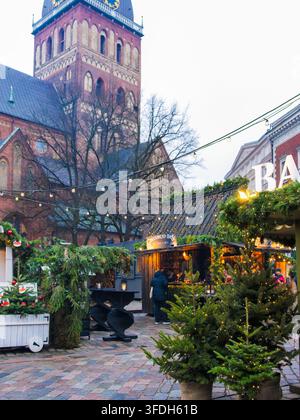 Die Kulisse des Heritage-Marktes verbindet immergrüne Stände und Tannenbäume mit einem hohen Backsteinturm einer Kirche in der Altstadt von Riga für die europäische Weihnachtstradition, Bogen Stockfoto