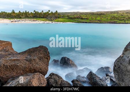 Blick auf den Anakena-Strand mit türkisfarbenem Wasser, vulkanischen Felsen im Vordergrund und Palmen entlang des sandigen Ufers auf der Osterinsel, Valparaíso, Chil Stockfoto