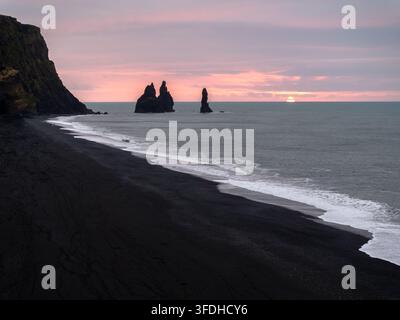 Luftbild des schwarzen Sandstrands von Reynisfjara mit den Reynisdrangar-Klippen, die aus dem Atlantischen Ozean unter einem weichen Sonnenuntergangshimmel in Island aufragen Stockfoto