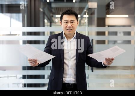 Asiatischer Geschäftsmann, der sich überfordert und gestresst fühlt, während er zwei Dokumente hält und mit einem komplexen Problem oder einer schwierigen Entscheidung in einer modernen Büroumgebung kämpft Stockfoto