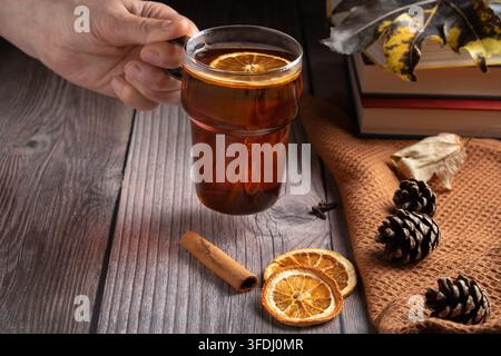Person hält eine warme Glastasse mit herbstgewürztem Tee und getrockneter Orange. Rustikale Holzeinrichtung mit Büchern und Kiefernzapfen Stockfoto