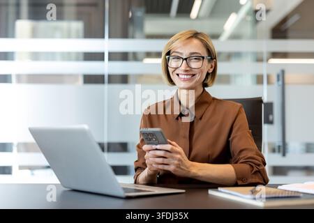 Selbstbewusste Geschäftsfrau lächelt in die Kamera, hält ein Smartphone und sitzt an ihrem Schreibtisch mit einem Laptop, was moderne Kommunikation und ein erfolgreiches Berufsleben widerspiegelt Stockfoto