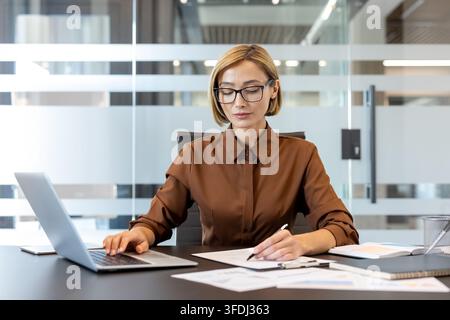 Professionelle Geschäftsfrau, die an ihrem modernen Bürotisch multitaskt, an einem Laptop arbeitet und gleichzeitig Notizen auf Dokumenten macht, konzentriert und effizient in einem Unternehmenskonto Stockfoto