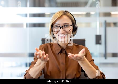 Zufriedene junge Geschäftsfrau trägt Headset und Brille, schaut in die Kamera, führt einen Videoanruf, kommuniziert mit Kunden, bietet Online-Kundensupport oder Beratung an Stockfoto