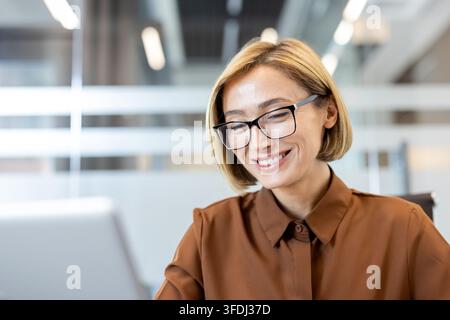 Professionelle blonde Frau mit Brille und braunem Hemd, die auf einen Laptop-Bildschirm hinunterschaut, glücklich lächelnd, während sie in einem hellen modernen Büroumfeld arbeitet Stockfoto