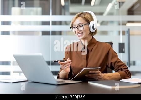 Geschäftsfrau mit Kopfhörern und Brille, die aktiv an einem Online-Meeting teilnimmt, Notizen macht, während sie lächelt und auf ihren Laptop-Bildschirm schaut, in einer modernen Büroumgebung Stockfoto