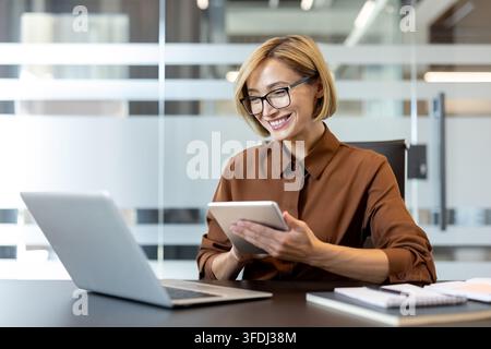 Professionelle Geschäftsfrau arbeitet in einem modernen Büro, glücklich mit ihrem Digitaltablet beschäftigt, während ein Laptop auf dem Schreibtisch liegt, und zeigt Multitasking und Technologieintegration in ihrer täglichen Arbeit Stockfoto