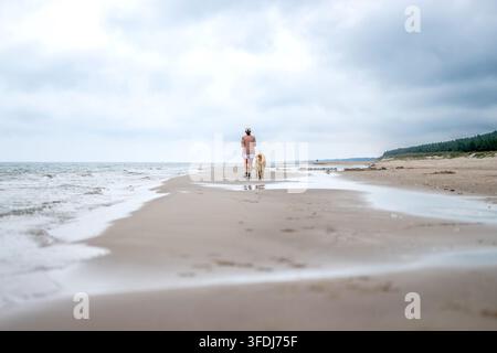 Frau geht an einem nassen Sandstrand mit goldenem Hund während eines bewölkten Morgens entlang Stockfoto