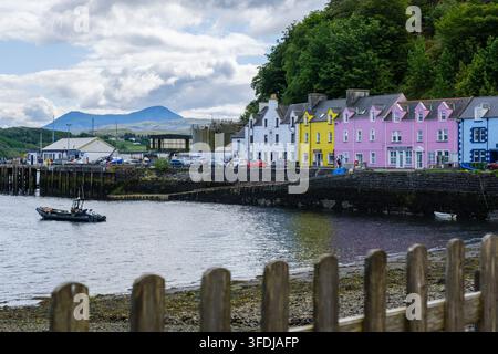 Hell bemalte weiße, gelbe, rosa und blaue Ufergebäude säumen den Hafen in Portree auf der Isle of Skye, Schottland, mit einem kleinen Boot Stockfoto