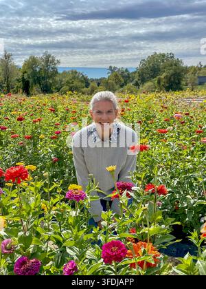 Eine lächelnde Frau in einem grauen Pullover steht inmitten eines lebendigen Feldes mit blühenden Zinnien in Rot-, Rosa-, Orange- und Gelbtönen, unter einem bewölkten Himmel und Tr Stockfoto