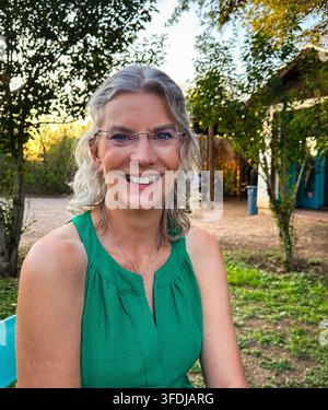 Eine fröhliche Frau mit lockigem grauen Haar und Brille lächelt herzlich im Freien in einer Garteneinstellung zur goldenen Stunde, trägt ein grünes Oberteil mit Lichterketten an Stockfoto