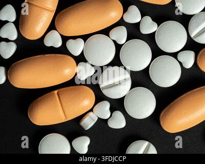 Verschiedene orangefarbene Kapseln und weiße runde und herzförmige Tabletten, die auf einem schwarzen Hintergrund verstreut sind, vermitteln Medizin, Gesundheitswesen, Liebe, Rezeptur, Stockfoto