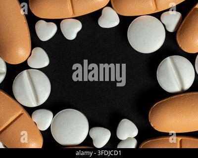 Verschiedene weiße und orangefarbene Tabletten, darunter kleine herzförmige Pillen, die als Bordüre auf einem schwarzen Hintergrund angeordnet sind, um Medikamente, Apotheke, Herz zu vermitteln Stockfoto