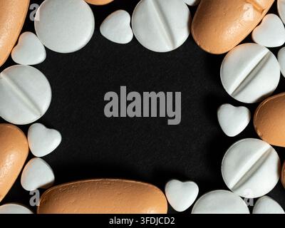 Herzförmige weiße Tabletten und runde orangefarbene sowie weiße Pillen, die um ein schwarzes Zentrum angeordnet sind und einen Rahmen für Medizin, Pharmazie und Gesundheitswesen bilden Stockfoto